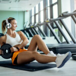 Smiling athletic woman using barbell disk while doing sit-ups during sports training in a gym.